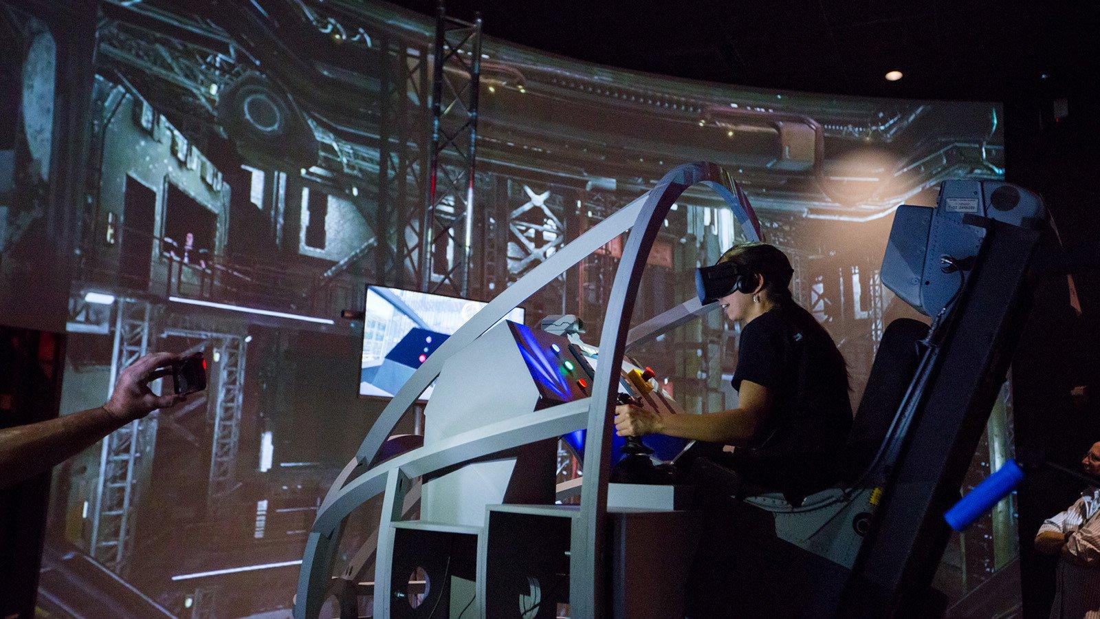一名戴着虚拟现实头盔的学生坐在航天飞机着陆模拟器中. 他们面前是一个展示仓库的大屏幕.