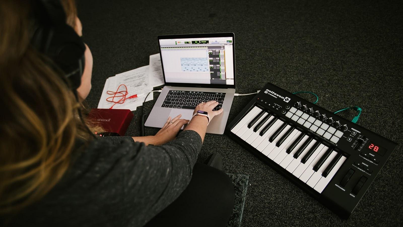 一个戴着耳机的学生坐在地板上，拿着笔记本电脑和电子键盘. 音乐编辑软件在笔记本电脑屏幕上.